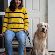 Woman wearing a striped yellow and black shirt sitting with golden retriever with a black leash.